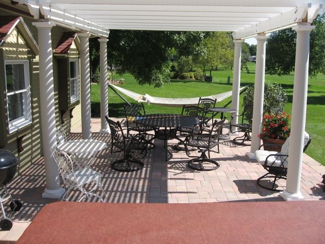 A white pergola covers a brick patio with an outdoor dining set, a hammock in the background, and a lawn beyond.
