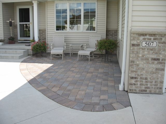 A patio with multi-toned stone pavers, two chairs, and a small table located in front of a house with beige siding.