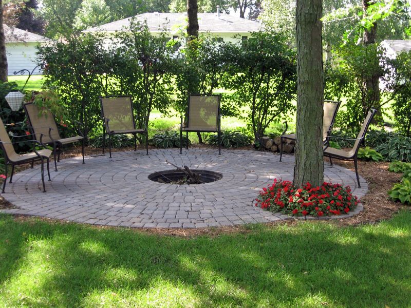 Circular stone patio with a central fire pit surrounded by outdoor chairs under a tree in a lush green backyard.
