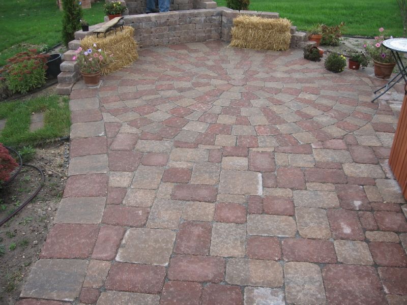 A circular patio made of multi-colored red and tan pavers, with a low stone retaining wall, potted plants, and hay bales.