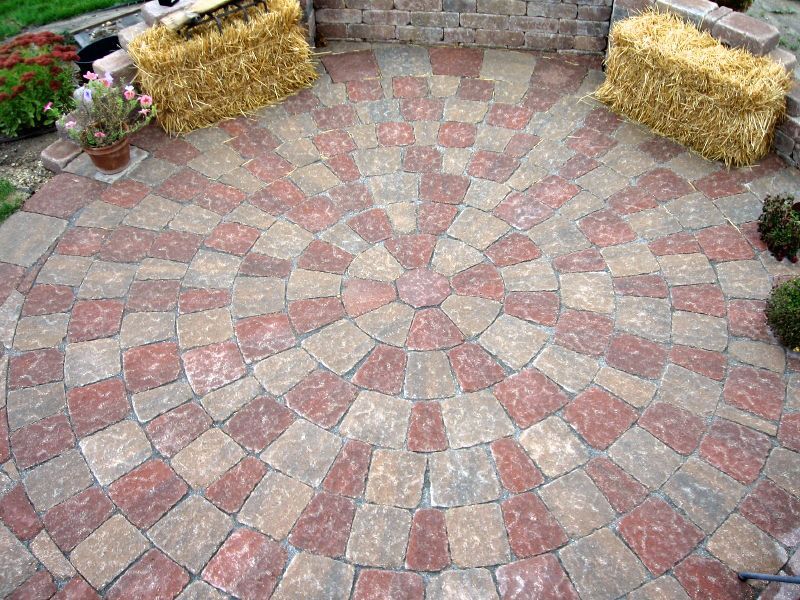 A circular patio made of alternating red and tan stone pavers, framed by a curved brick wall and two straw bales.