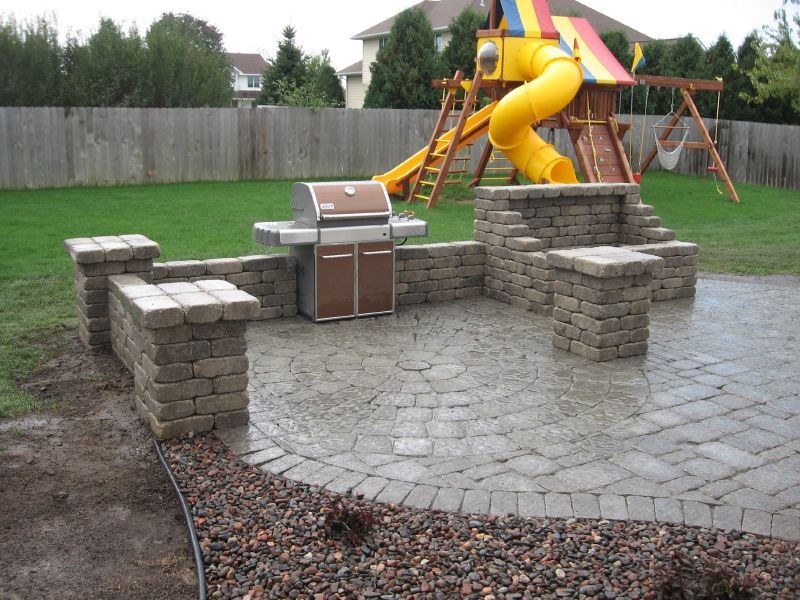 A backyard patio features a stone-built grill station and a circular paver floor, with a colorful play set in the distance.