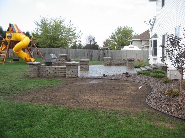A stone patio with a built-in grill and retaining walls in a backyard, beside a house and a yellow playground set.