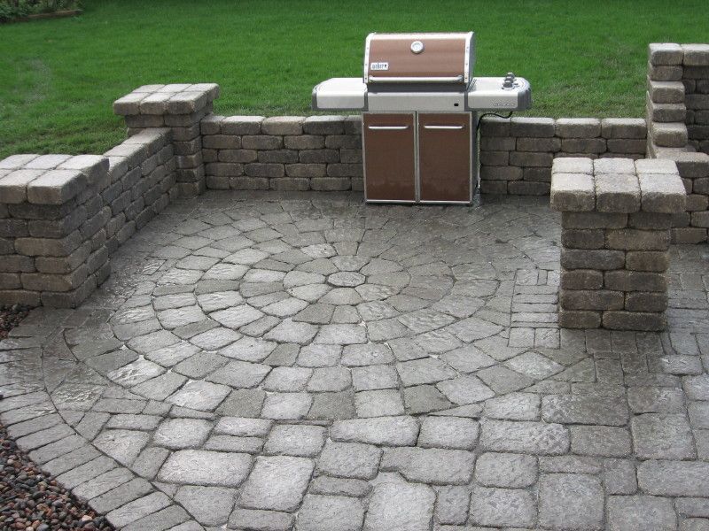 A stone patio featuring a circular paver pattern, a built-in grill, and low stone retaining walls on a grassy lawn.
