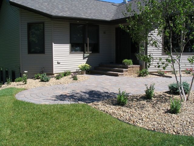 A suburban house with light siding and a circular paver patio surrounded by gravel landscaping and green lawn.