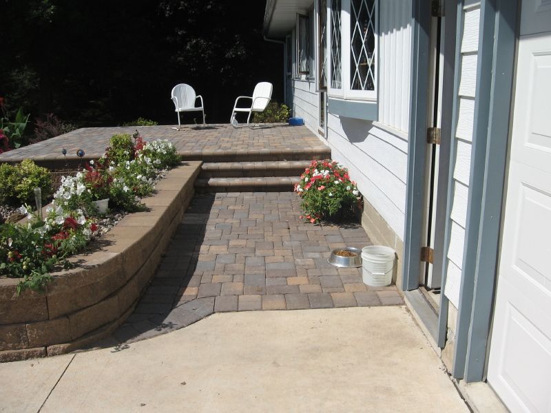 A stone-paved walkway leads to two steps and a patio with white chairs beside a house with a retaining wall and flowers.