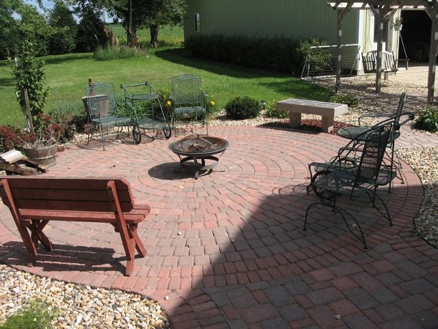A circular red brick patio with a fire pit, surrounded by various benches and chairs in a grassy backyard setting.