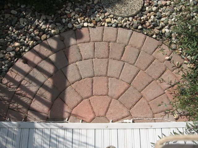A semi-circular stone patio made of tan and reddish-brown pavers, bordering a wooden deck and a bed of grey river rocks.