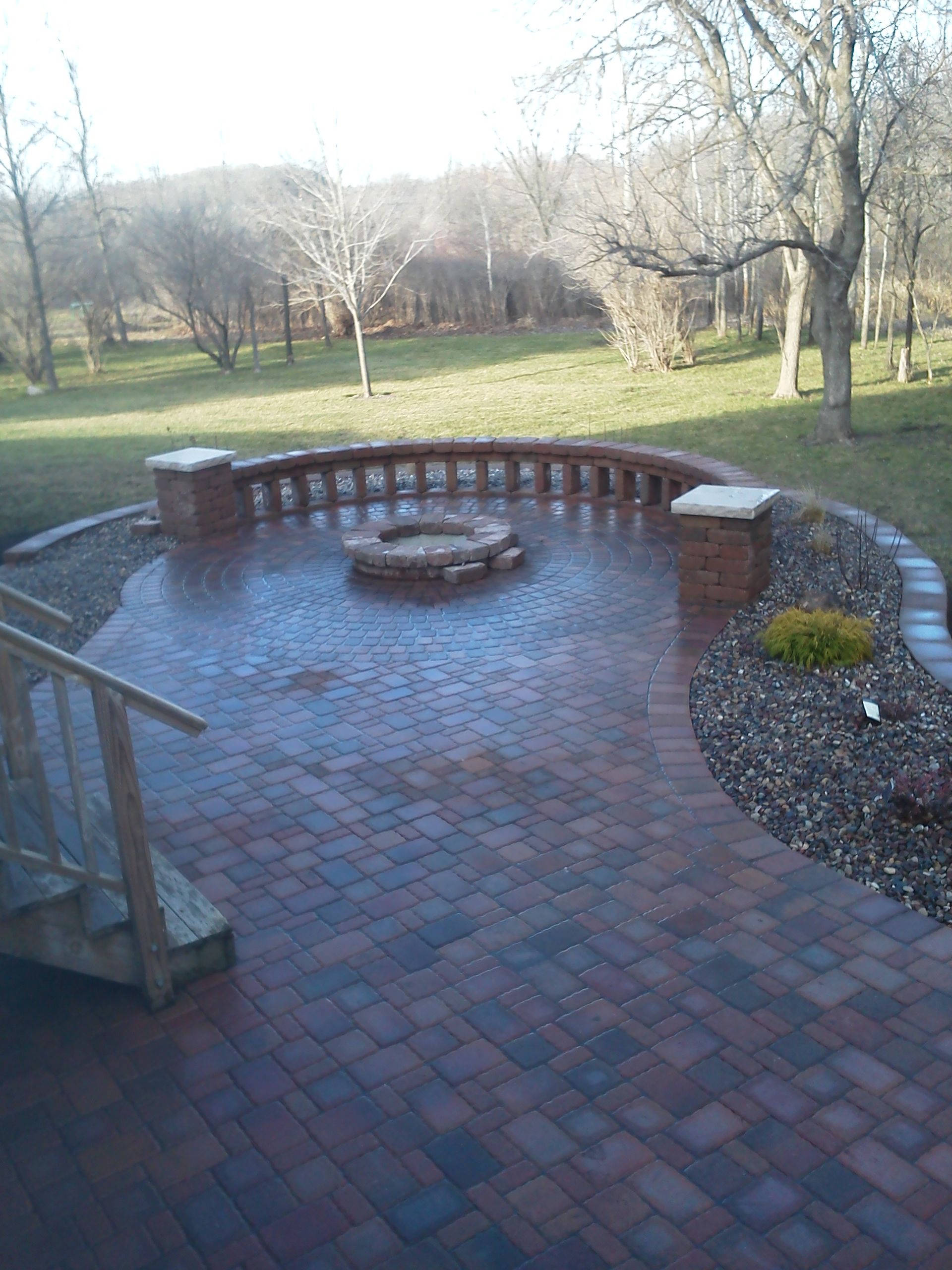 A red-brick patio featuring a central circular fire pit and a curved brick bench, surrounded by a grassy yard and trees.