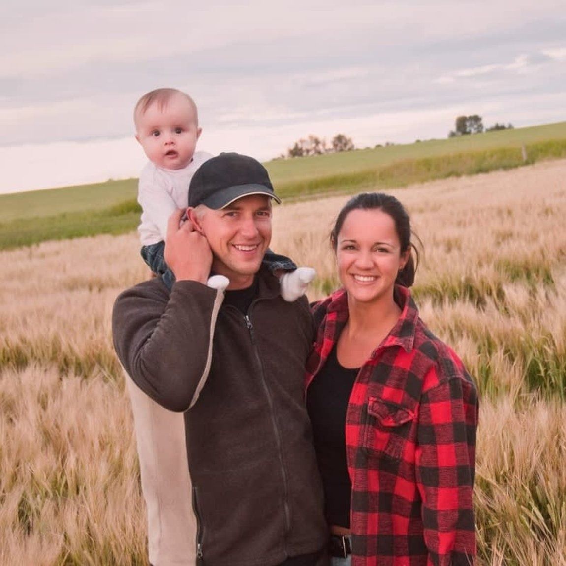 Family of three smiles in a wheat field; father carries baby on shoulders, mother smiles beside.