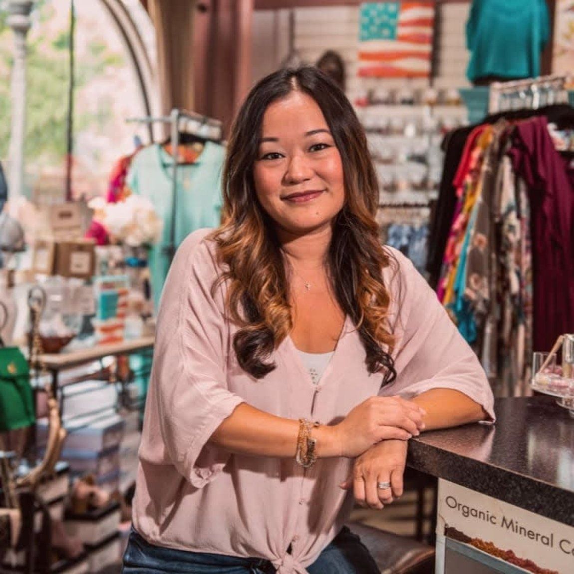 Woman smiling, leaning on counter in a retail store with clothing and merchandise visible.