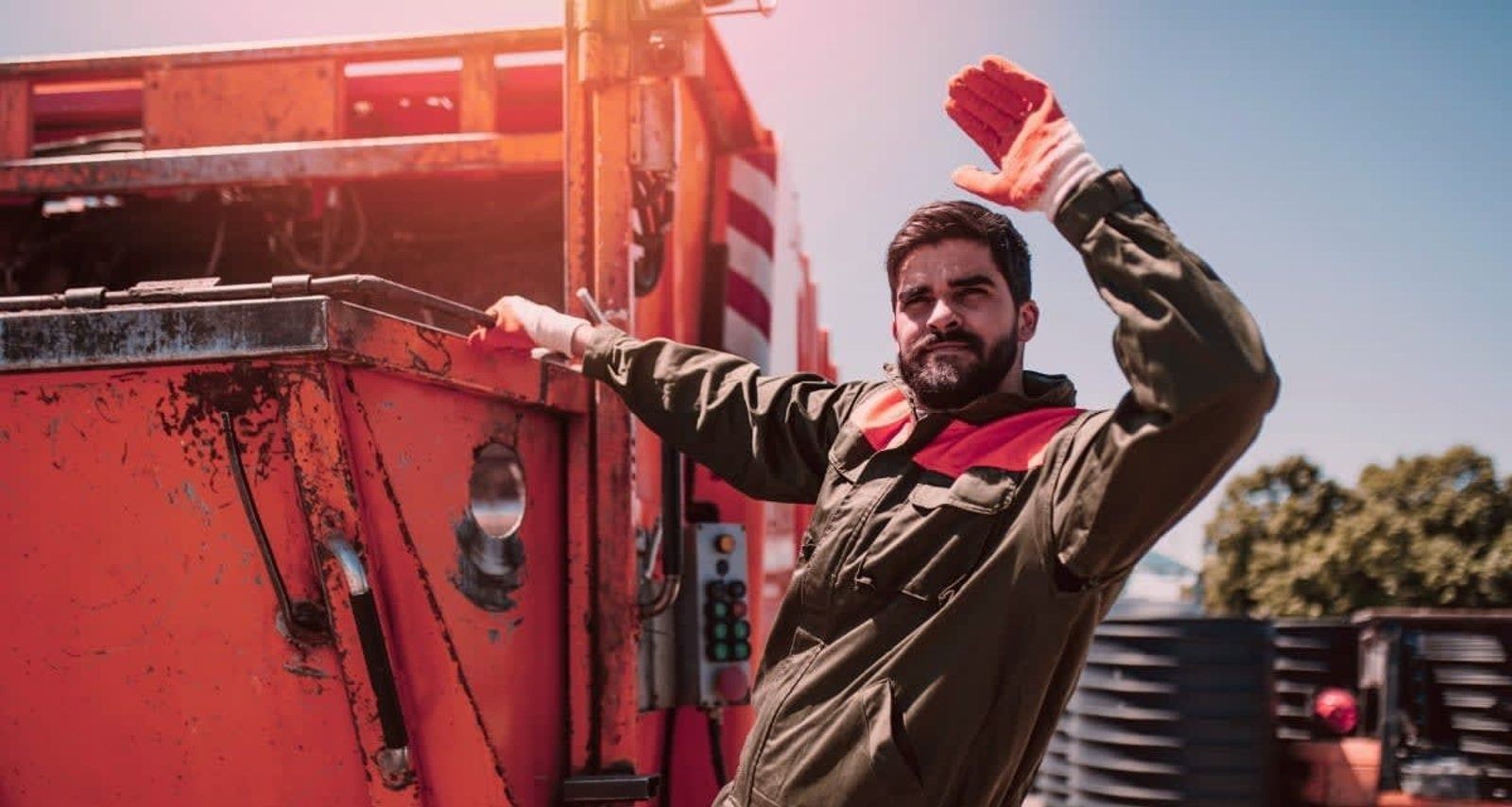Garbage truck worker waving with red-gloved hand near orange truck, sunny outdoor scene.