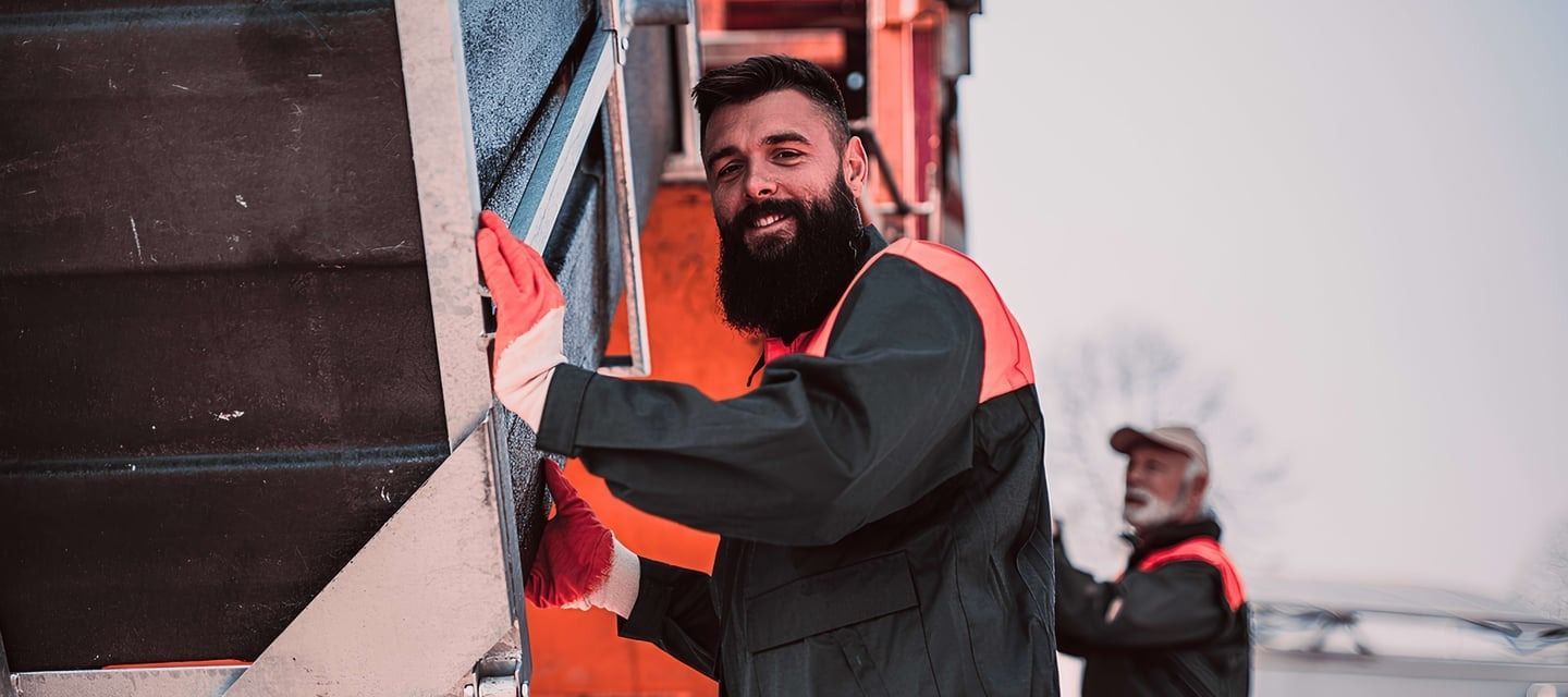 Man with a beard in orange and black work clothes holding onto stairs, smiling. Another person visible in the background.