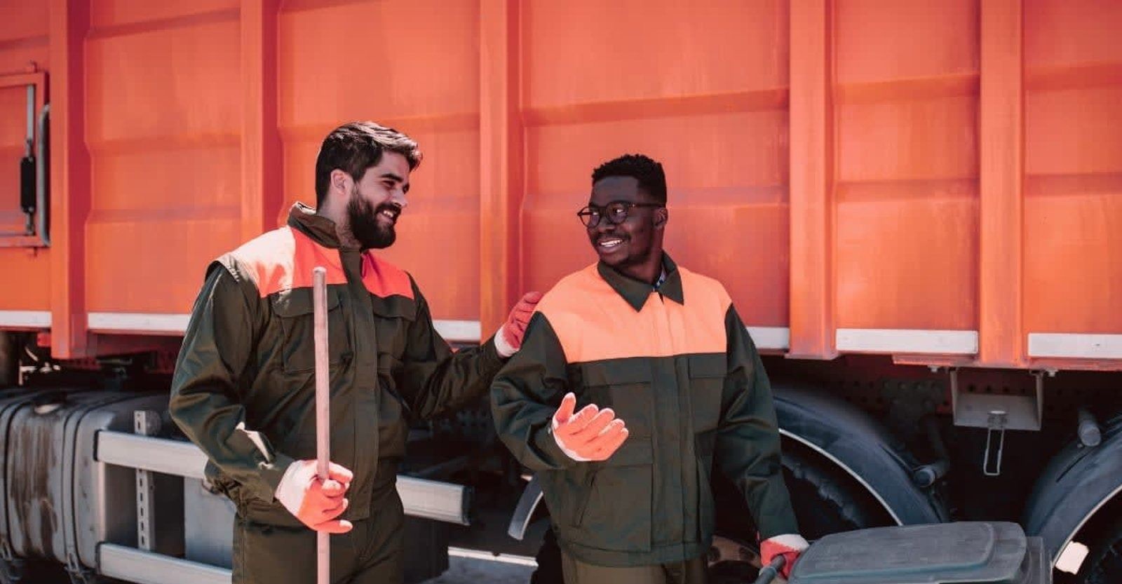 Two sanitation workers smiling near an orange truck. One holds a broom.