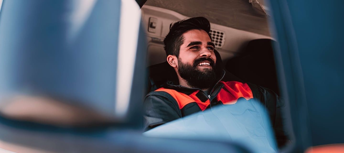Man with a beard smiling inside a vehicle, possibly a truck, wearing a safety vest.