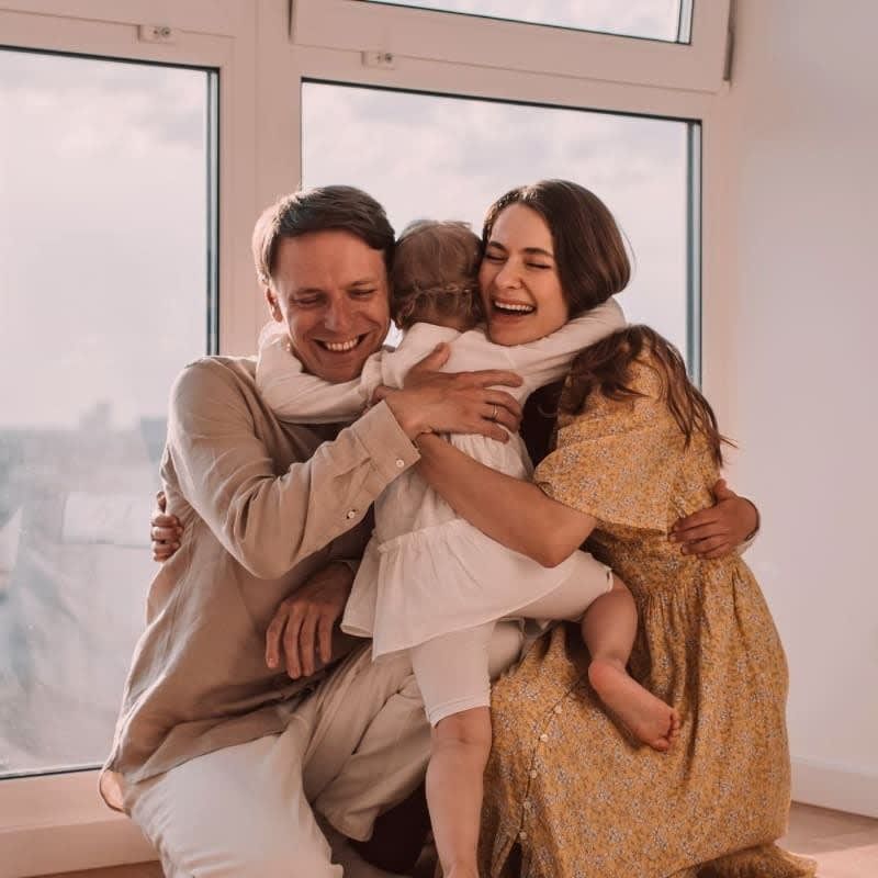 Family hugging near a window: Two adults and two children embracing, smiling with joy. Sunlight illuminates them.
