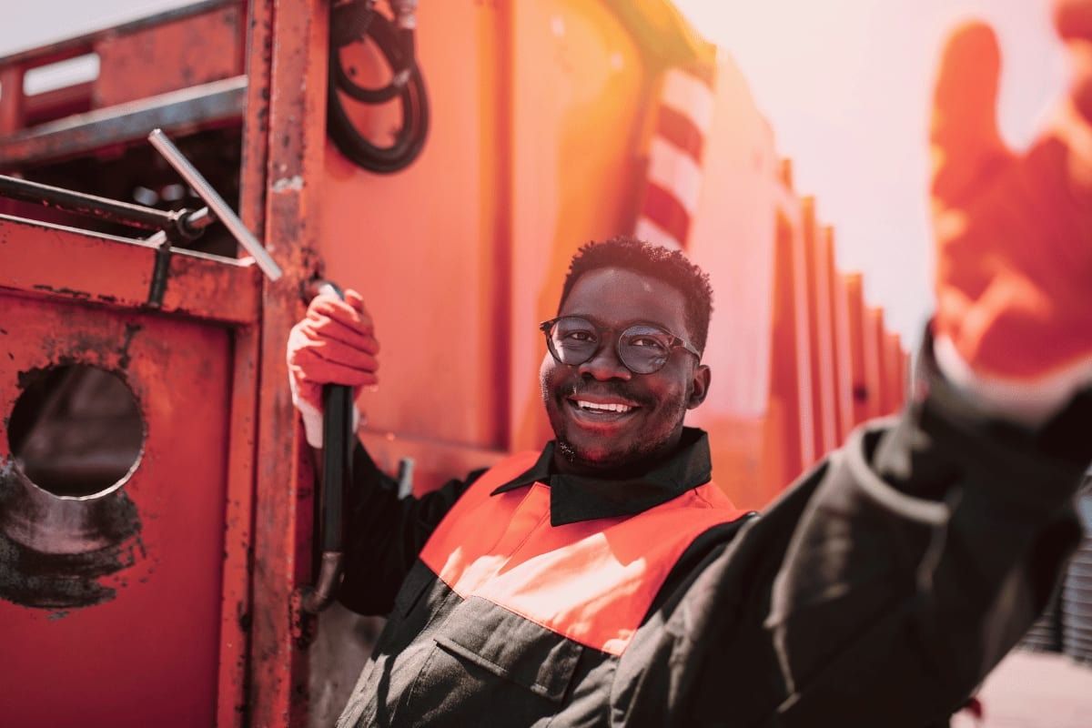 Garbage truck worker smiling, wearing orange gloves and vest, holding a tool. Bright, sunny day.