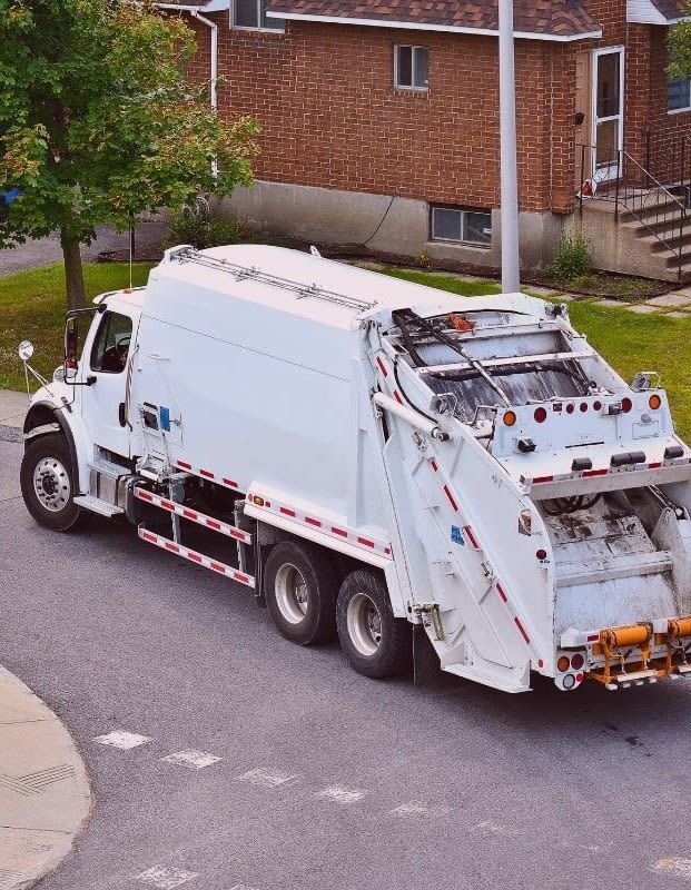 White garbage truck driving down a street in front of a brick house.