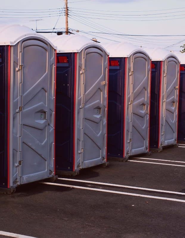 Line of portable toilets in a parking lot; gray and blue plastic with white and red accents.