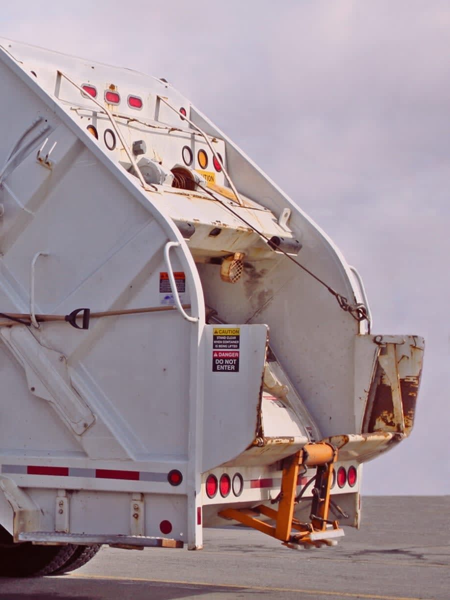 Rear view of a white garbage truck with open container, red lights, and a yellow lift arm.