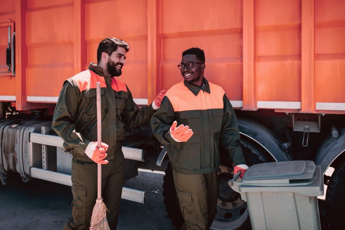 Two sanitation workers smiling near an orange garbage truck; one holds a broom, the other a trash bin.