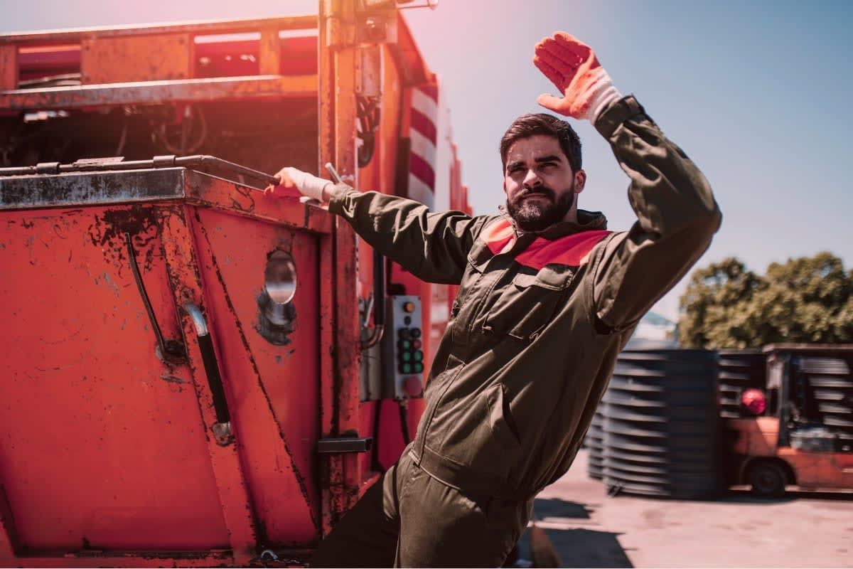 Man in work clothes near an orange truck, raising a gloved hand. Outdoor setting with forklift.