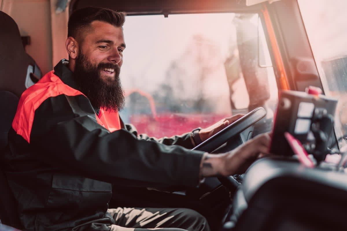 Man with beard smiles while driving a vehicle, wearing a safety vest.