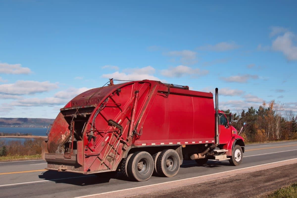 Red garbage truck driving on a road, blue sky background.