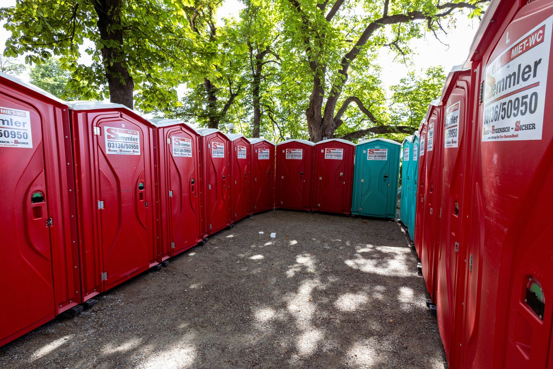 Rows of red portable toilets along a gravel path under green trees, with one teal unit at the end