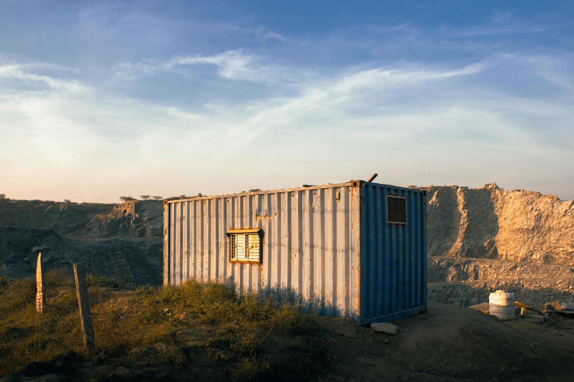 Blue shipping container with small window sits in a quarry under a blue sky.