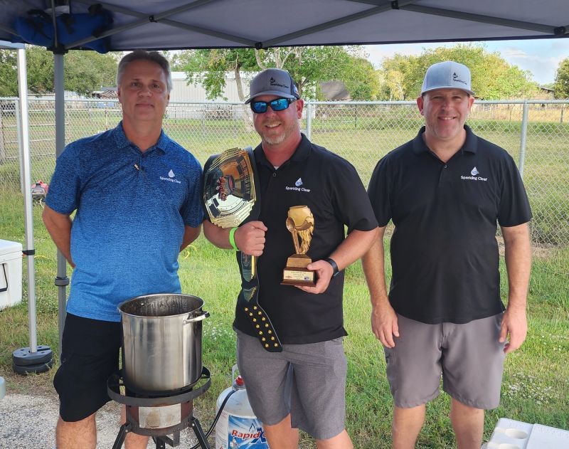 Three men under a canopy, one holding a trophy and belt, posing next to a pot of chili, outdoor setting.