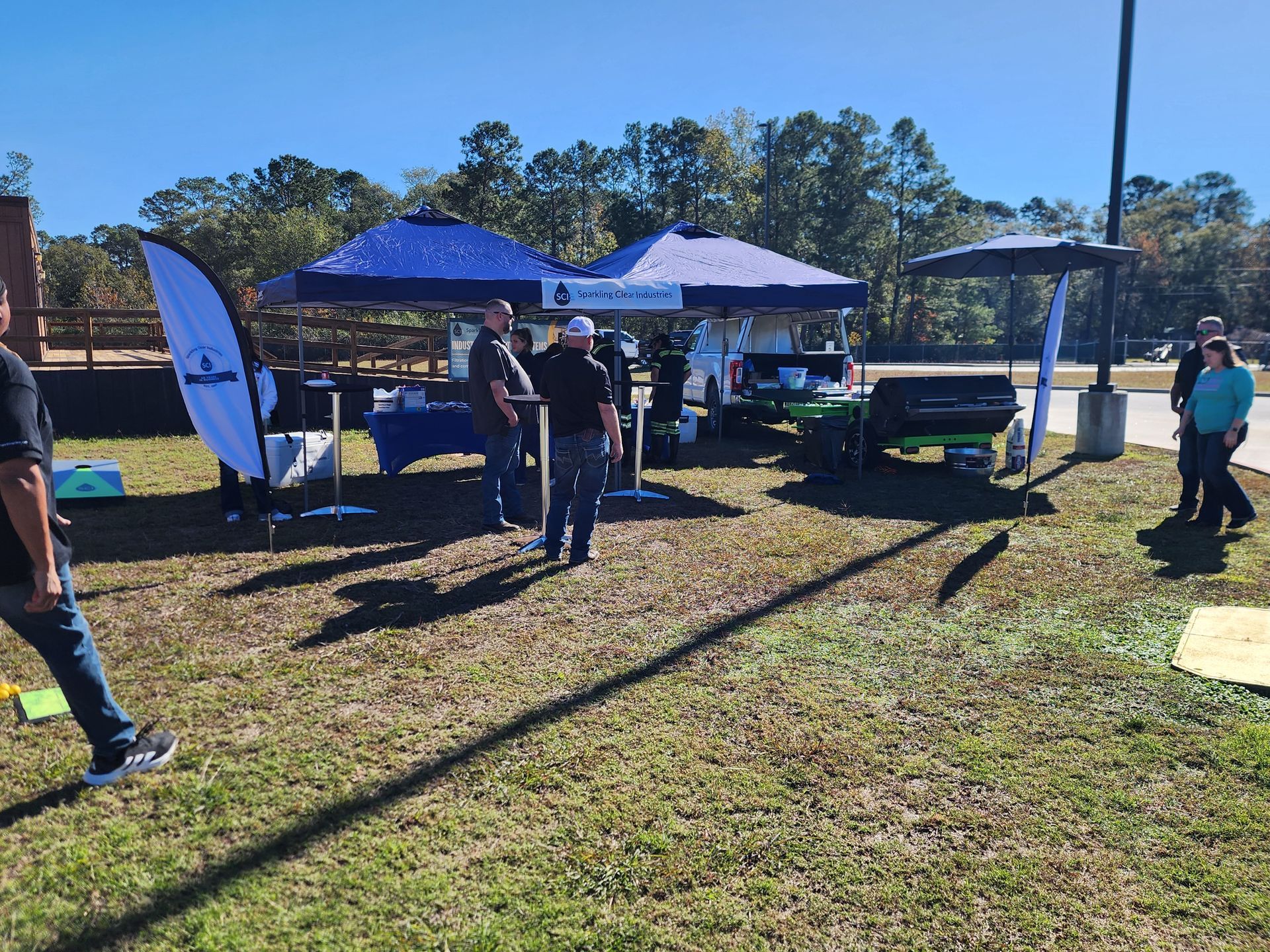 Outdoor event with blue tents, tables, and people interacting on a grassy area under a sunny sky.