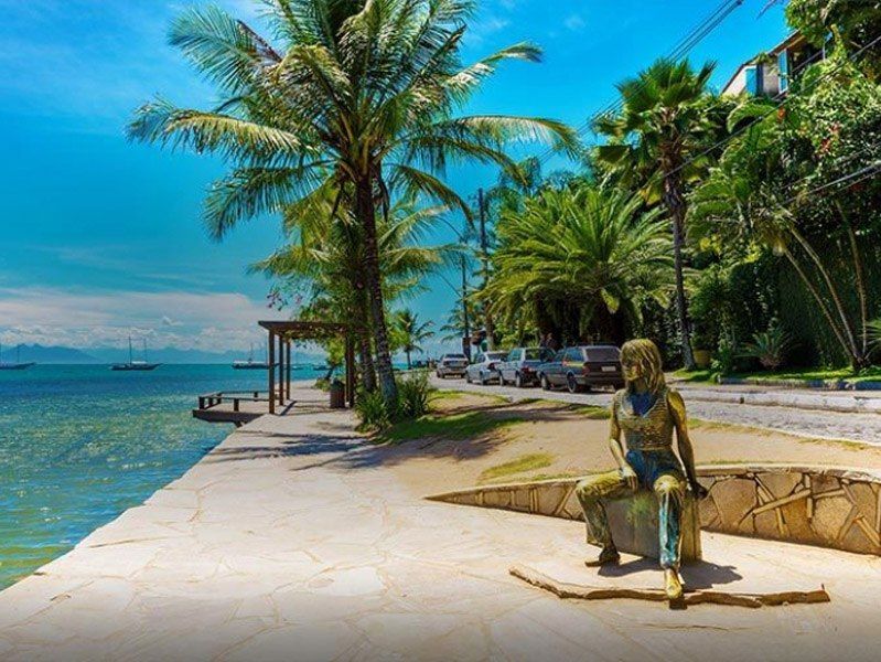 Calçadão à beira-mar com uma estátua de uma pessoa, palmeiras, rua com carros e oceano azul.