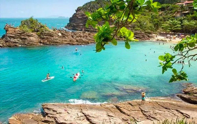 Baía turquesa com pessoas praticando stand-up paddle e nadando. Penhascos cobertos de vegetação emolduram a paisagem.