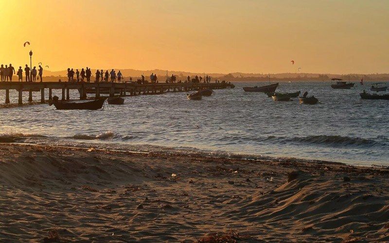 Pôr do sol sobre o oceano, píer com pessoas, barcos de pesca na água, praia de areia em primeiro plano.