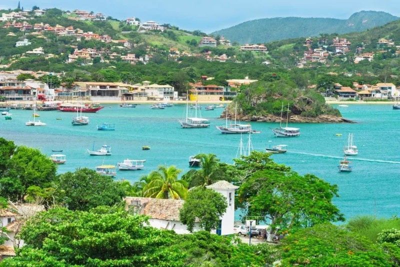 Vista panorâmica da costa com água turquesa, veleiros e colinas verdejantes pontilhadas de edifícios.