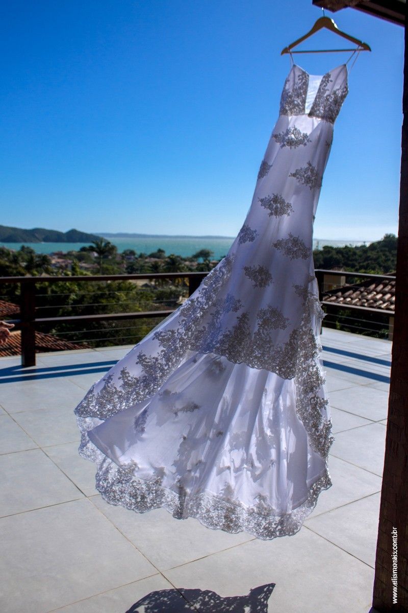 Vestido de noiva branco com bordados prateados pendurado em um cabide de madeira ao ar livre, com uma paisagem ao fundo.