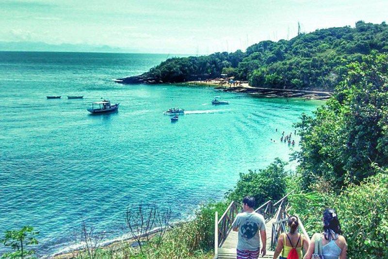 Pessoas descem degraus em direção a uma baía turquesa com barcos e árvores verdejantes.