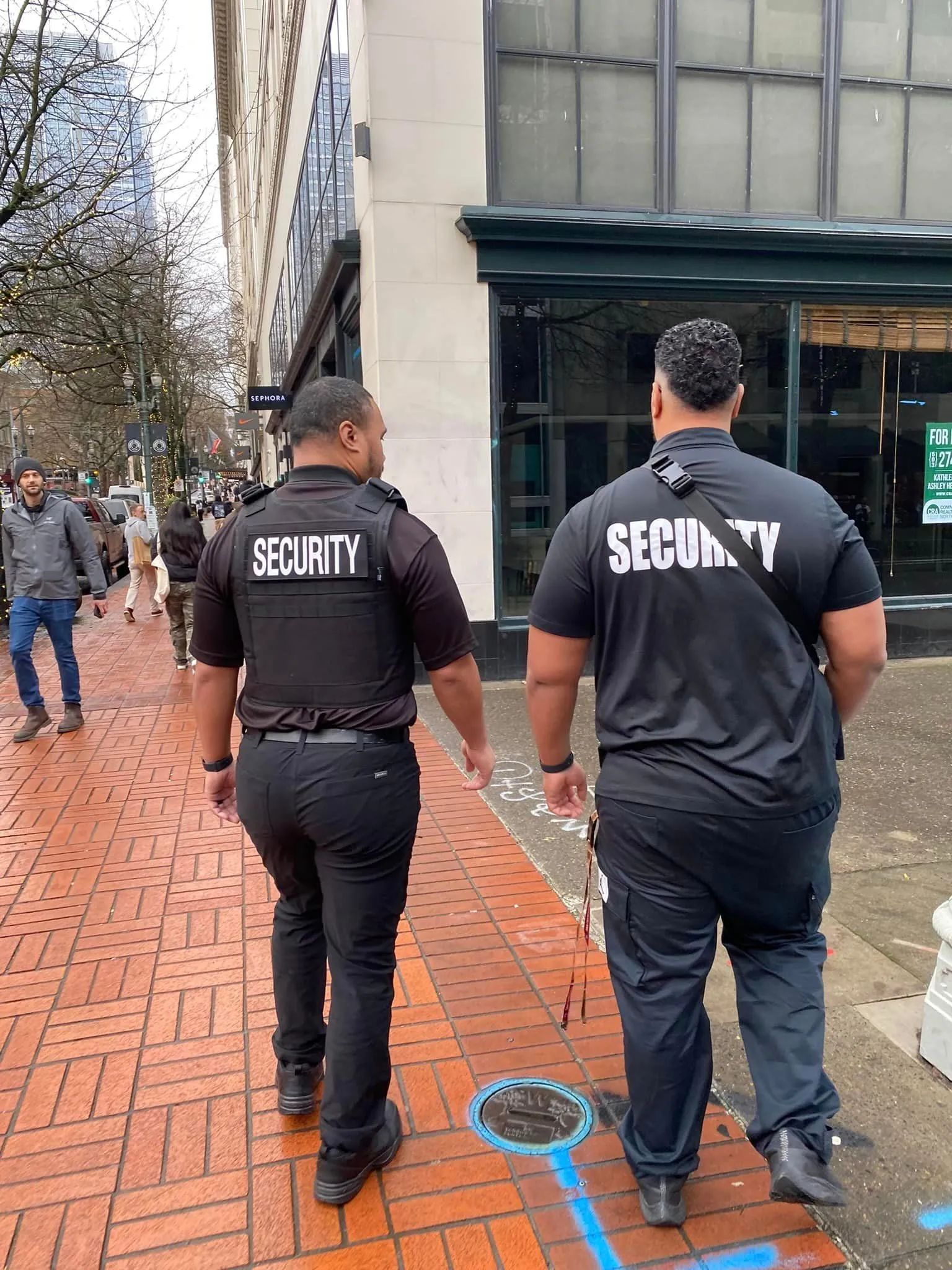 Two security guards in black shirts walk along a brick sidewalk beside a building entrance.