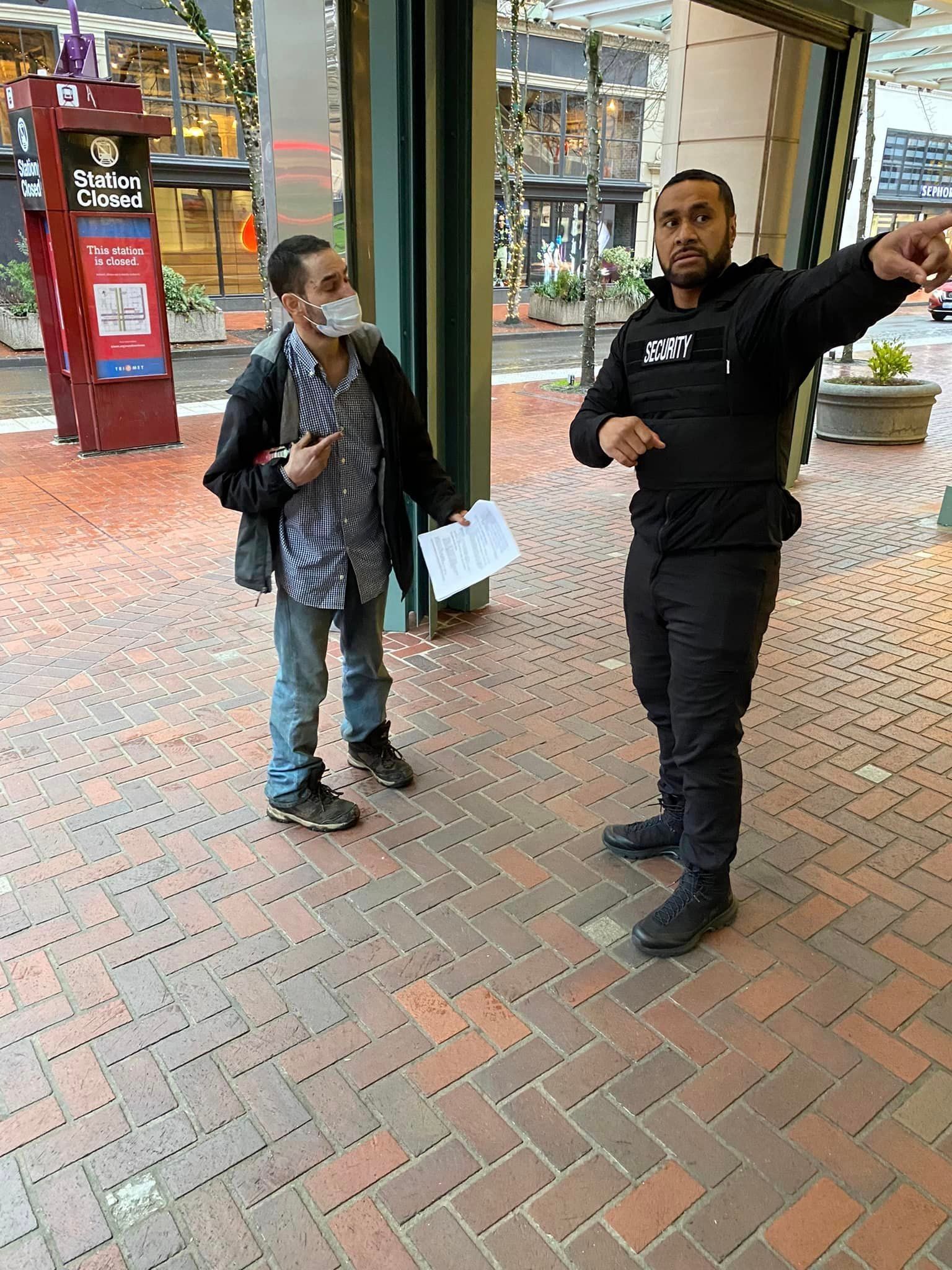 A man wearing a mask is standing next to another man on a brick sidewalk.