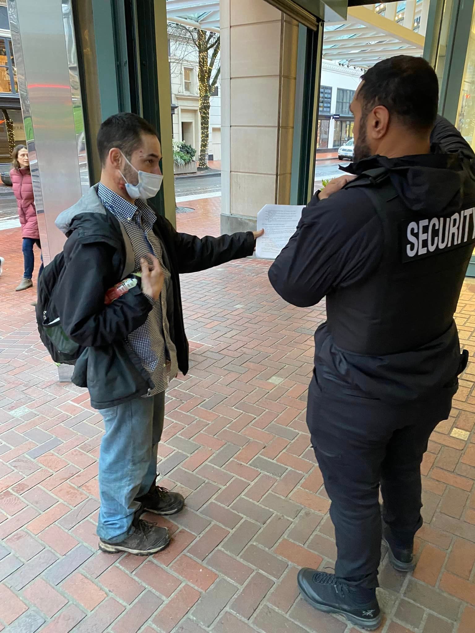 A man wearing a mask is talking to a security guard on a sidewalk.