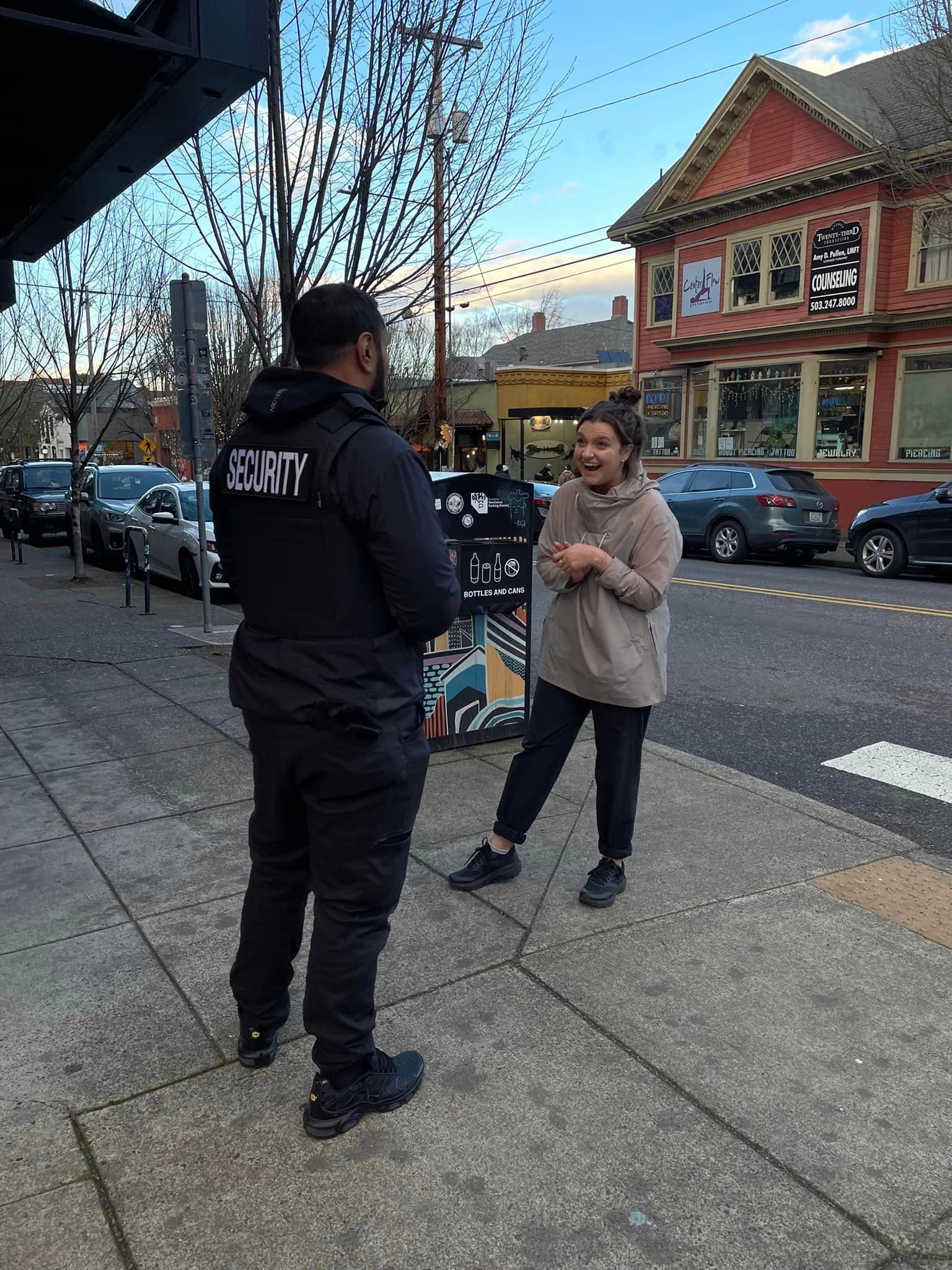 A man in a black jacket is talking to a woman on the sidewalk.