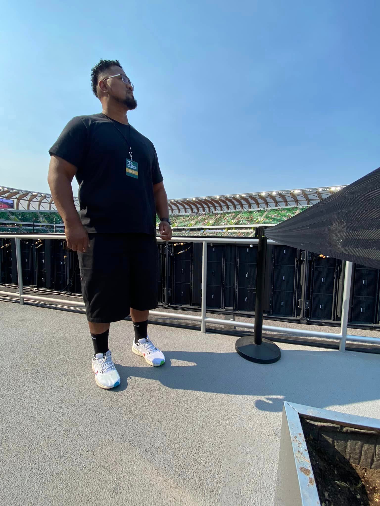 A man in a black shirt and shorts is standing in front of a fence.