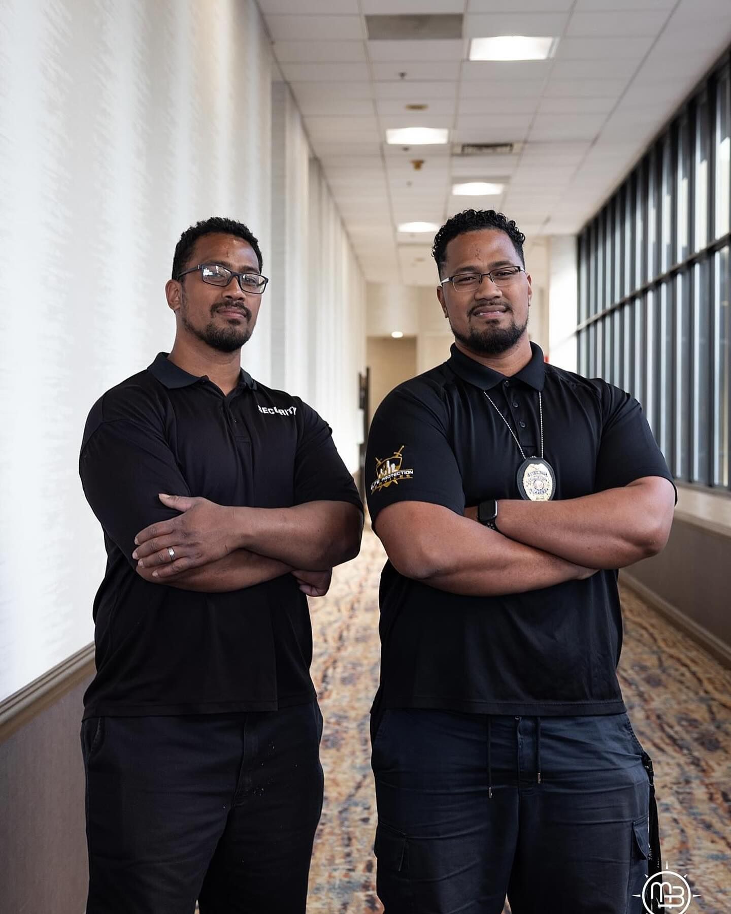 Two men are standing next to each other in a hallway with their arms crossed.