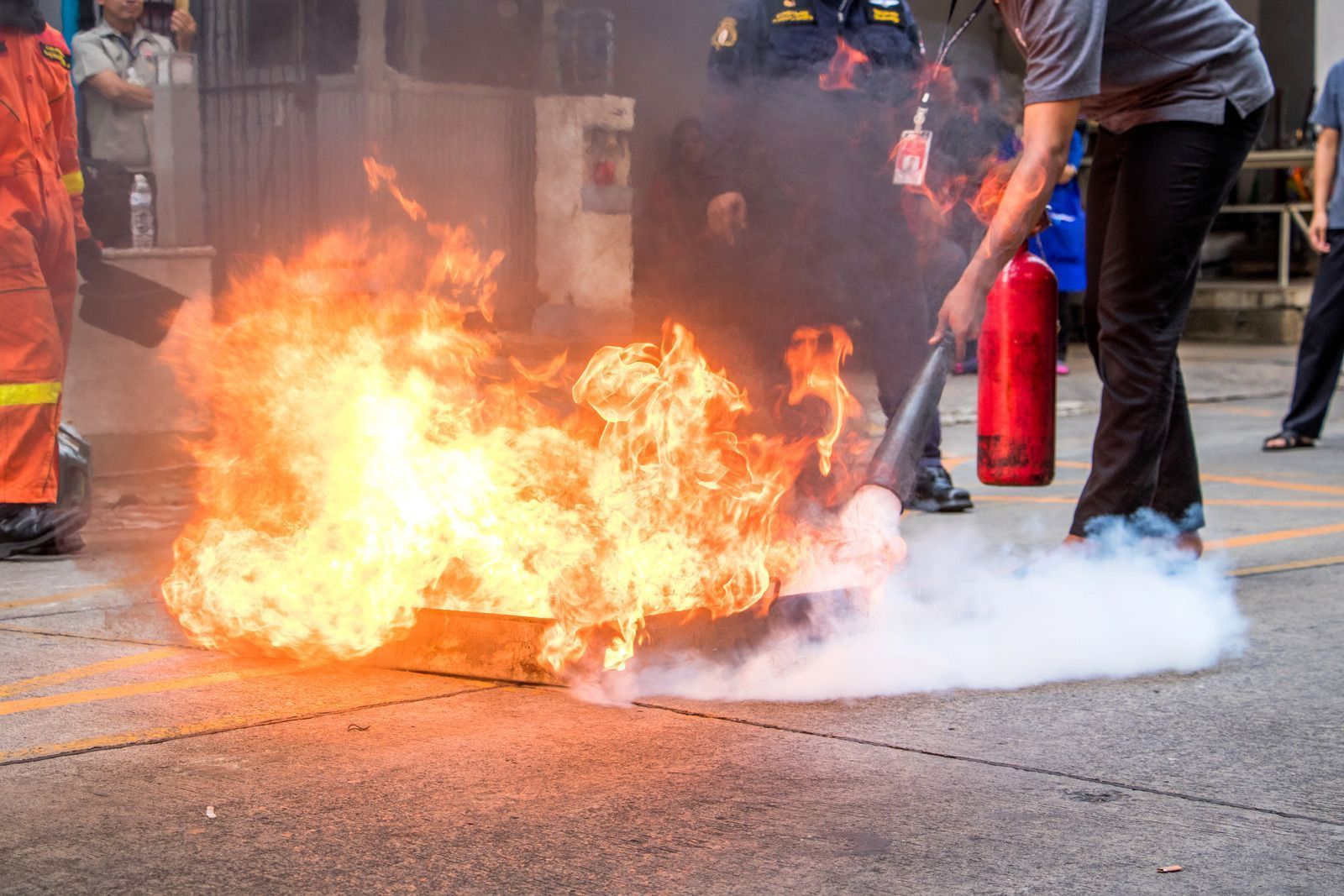 Person using a fire extinguisher on a small fire outdoors; flames and smoke visible.