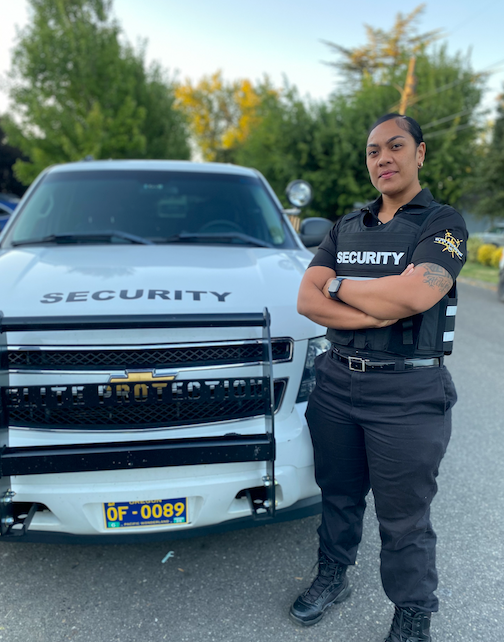 Woman security guard stands by white security vehicle, arms crossed, in front of a building.