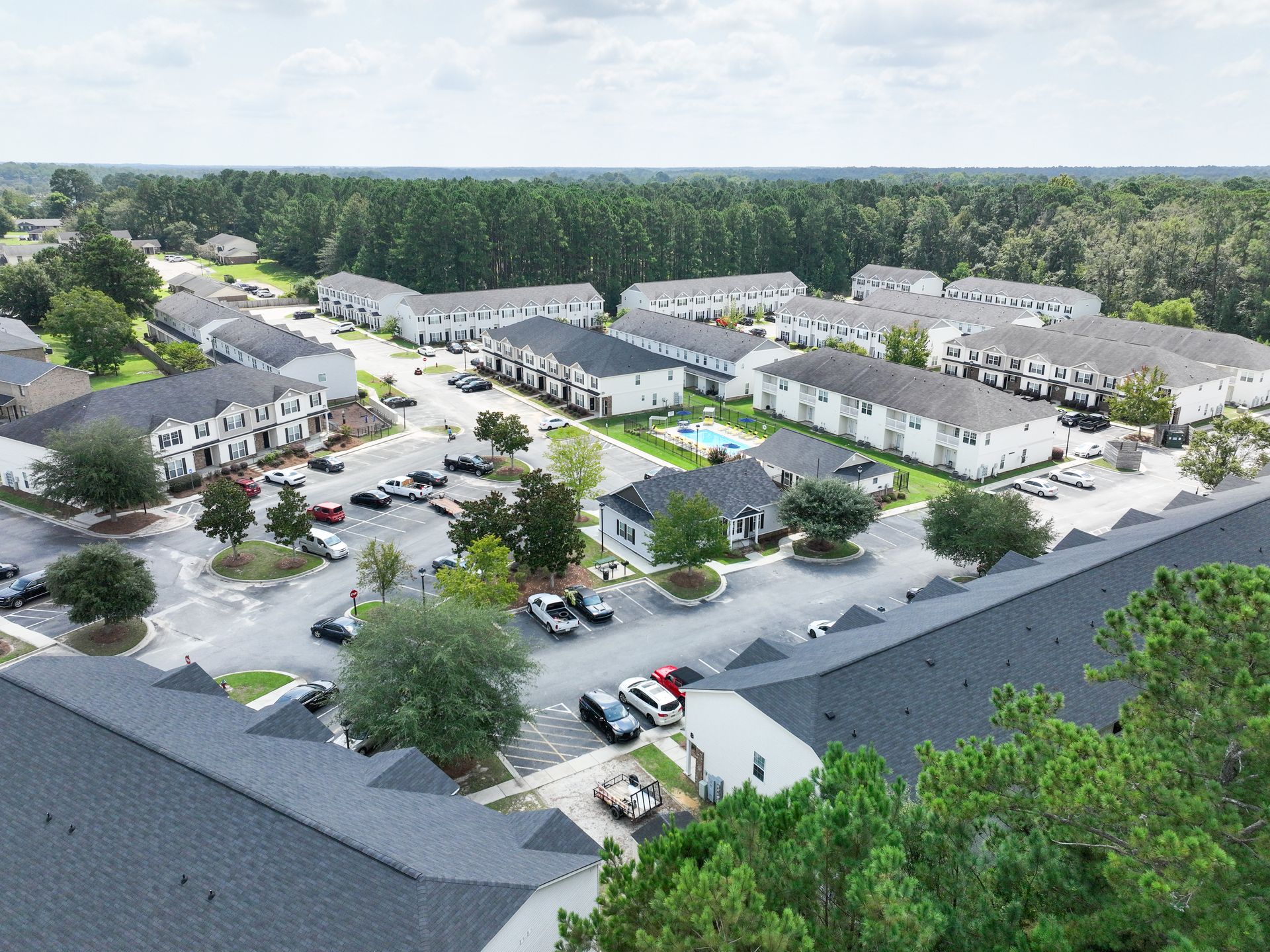Aerial view of a residential complex with white buildings, parking, a pool, and green trees under a blue sky.