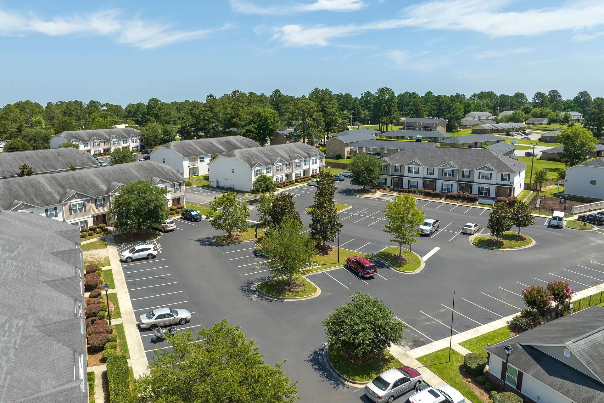 Aerial view of an apartment complex with parking lot and trees under a blue sky.