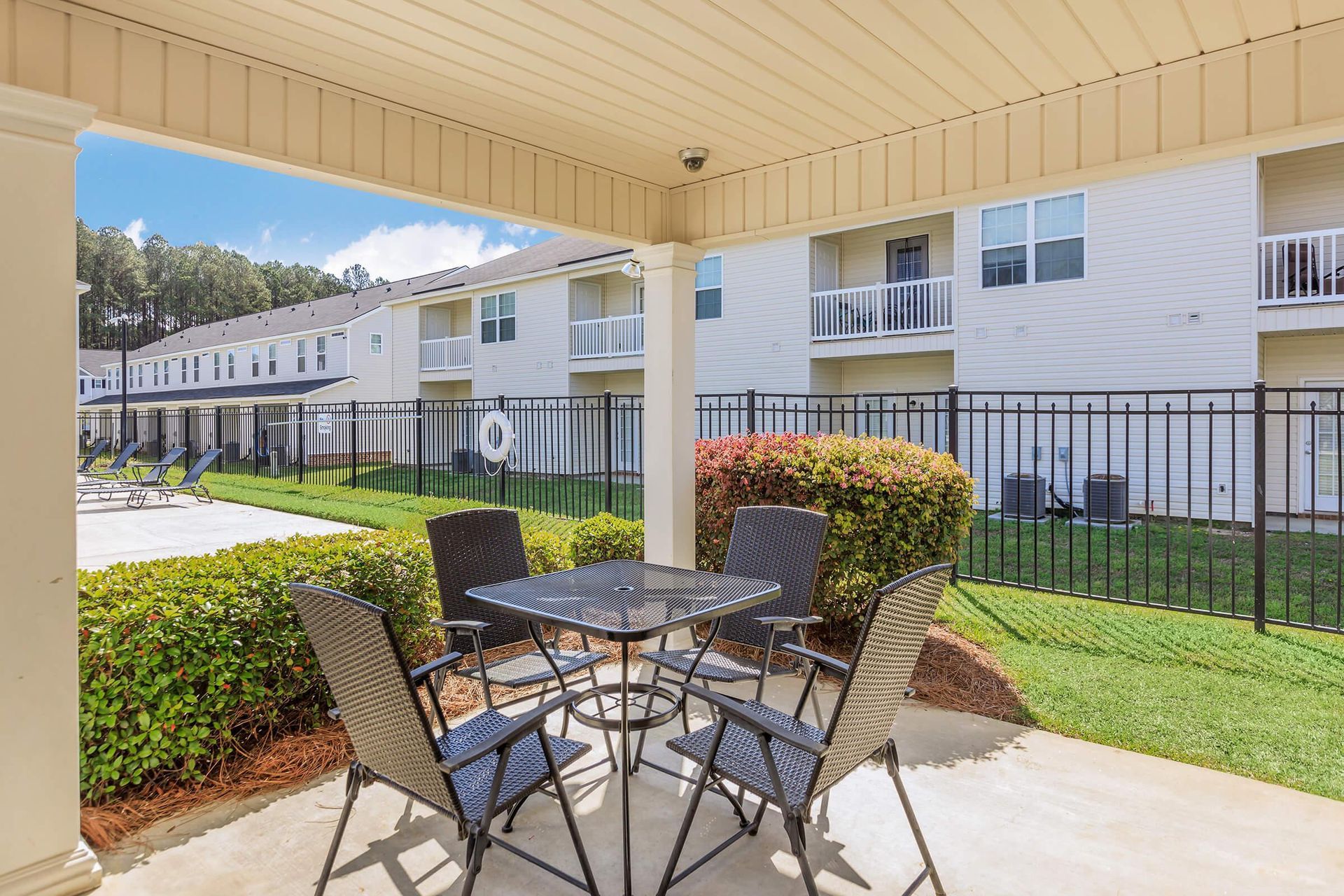 Patio with table and chairs, overlooking apartments. Green bushes and fence line the edge.
