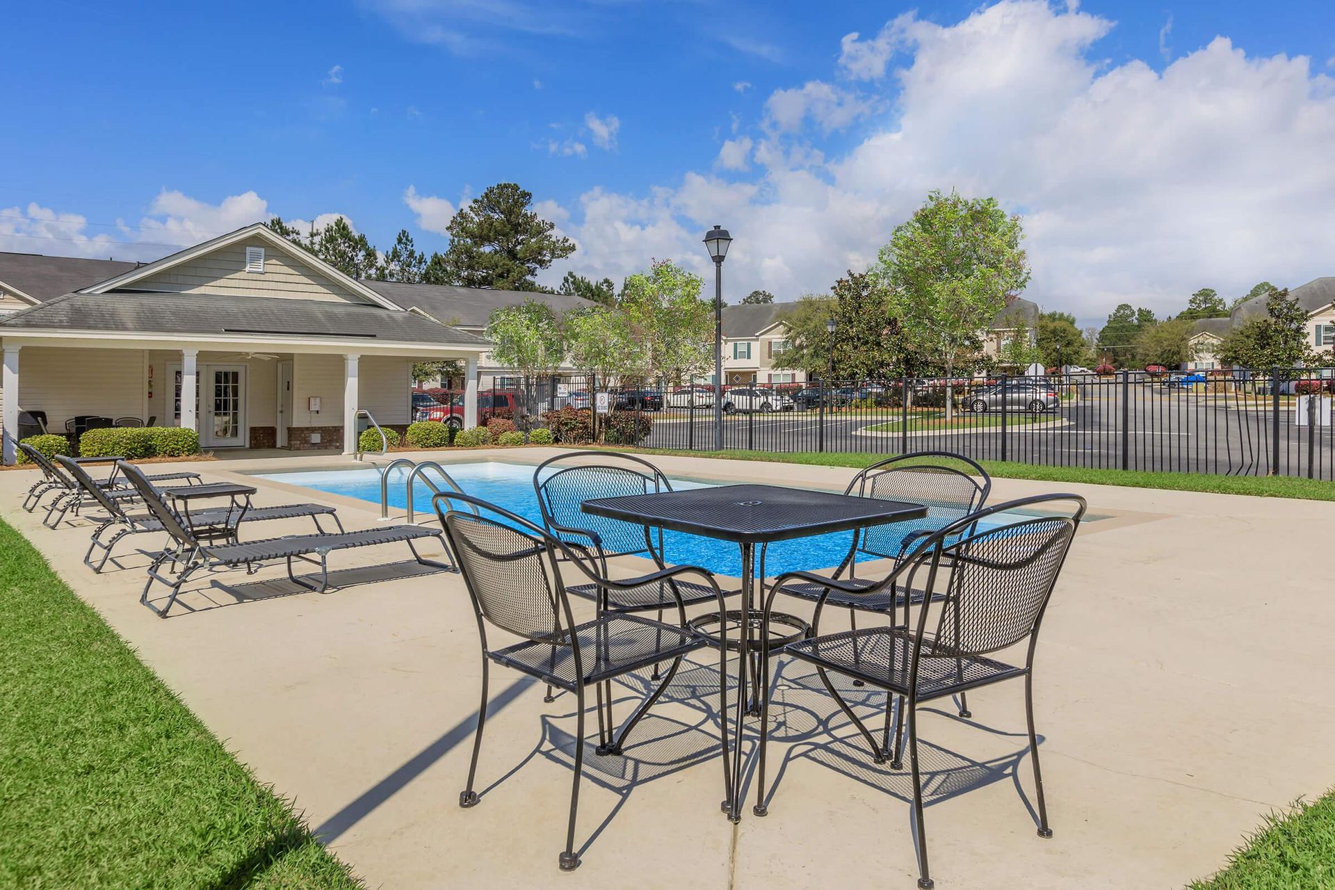 Poolside area with black metal table/chairs, lounge chairs, and building under blue sky.